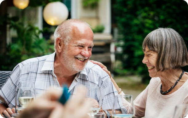 An older couple sit at an outdoor dinner table, smiling and laughing with each other. 