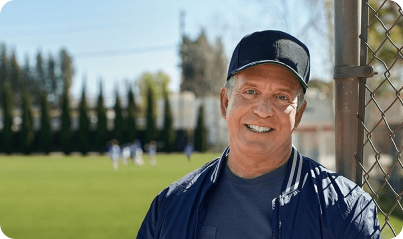 A baseball coach poses outside of a baseball diamond on a bright, sunny day.