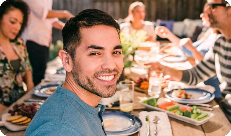 A man wearing a hearing aid smiles over his shoulder while enjoying an outdoor dinner with friends.