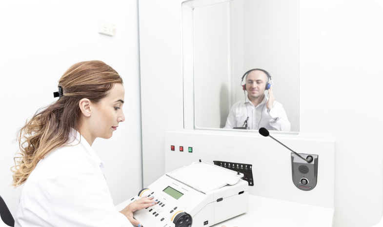 A Beltone technician performs a hearing test with a patient. A man sits in a sound booth, behind a window, while the audiologist performs the hearing test.