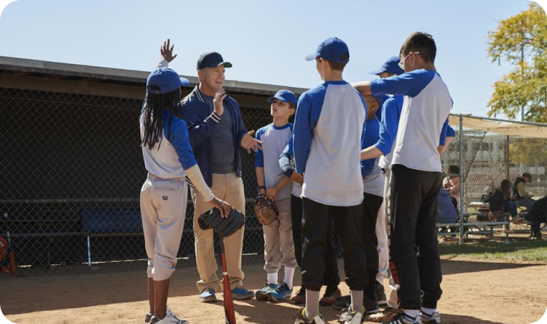 A baseball coach stands with a group of youth baseball players outside of a dugout, about to play a baseball game.