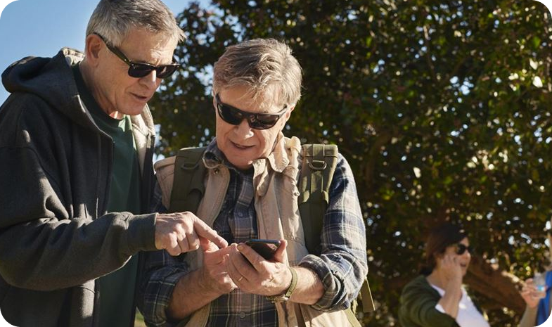 Two older adults look at a smartphone while outdoors in the sunshine.