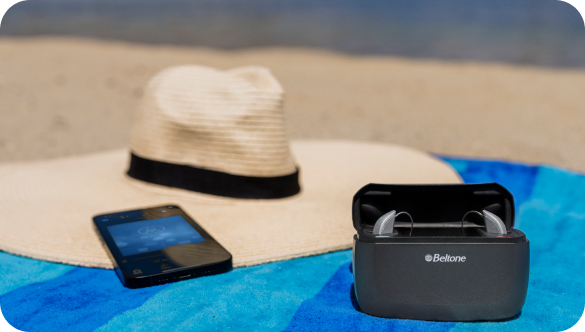The Beltone Achieve hearing aid is pictured sitting next to a smartphone and sun hat, on a beach towel.
