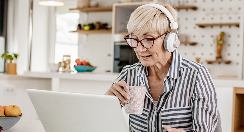 An older woman sits at a laptop, wearing headphones, and drinking from a mug of coffee. She is about to take a free online hearing test.