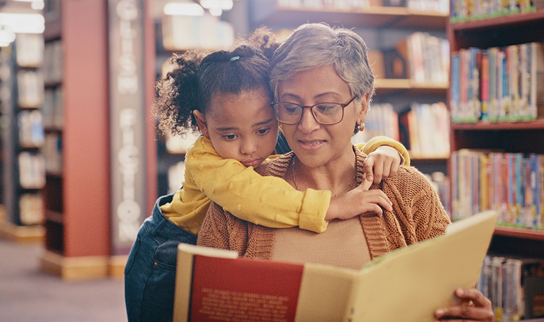 A young child reads a book in a library with her grandmother.