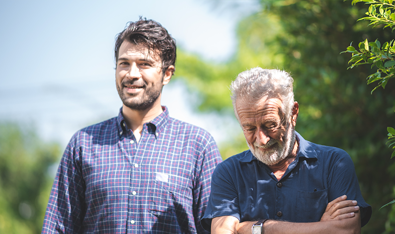 A younger man and older man stand next to each other while outside on a walk. The older man has his arms crossed and is looking at the ground, while the younger man smiles at the camera.