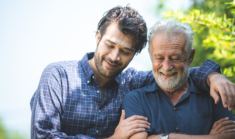 A younger man stands with his arm around an older man's shoulders.