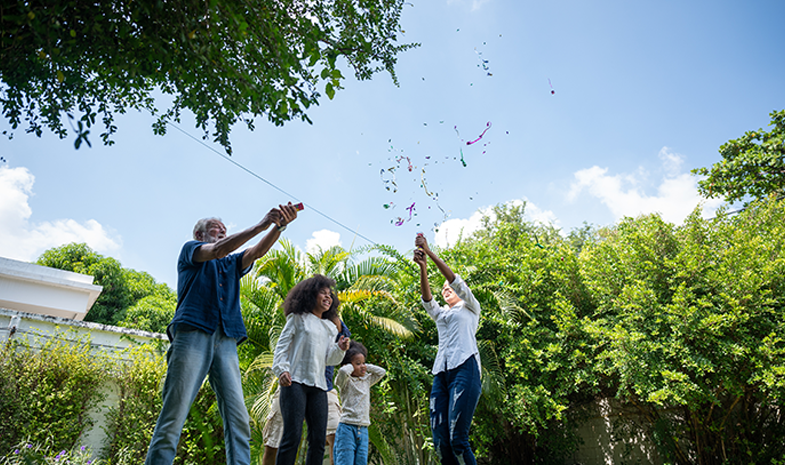 A family spends time together outside in a lush backyard. An older man, young woman, and two kids toss leaves up in the air.